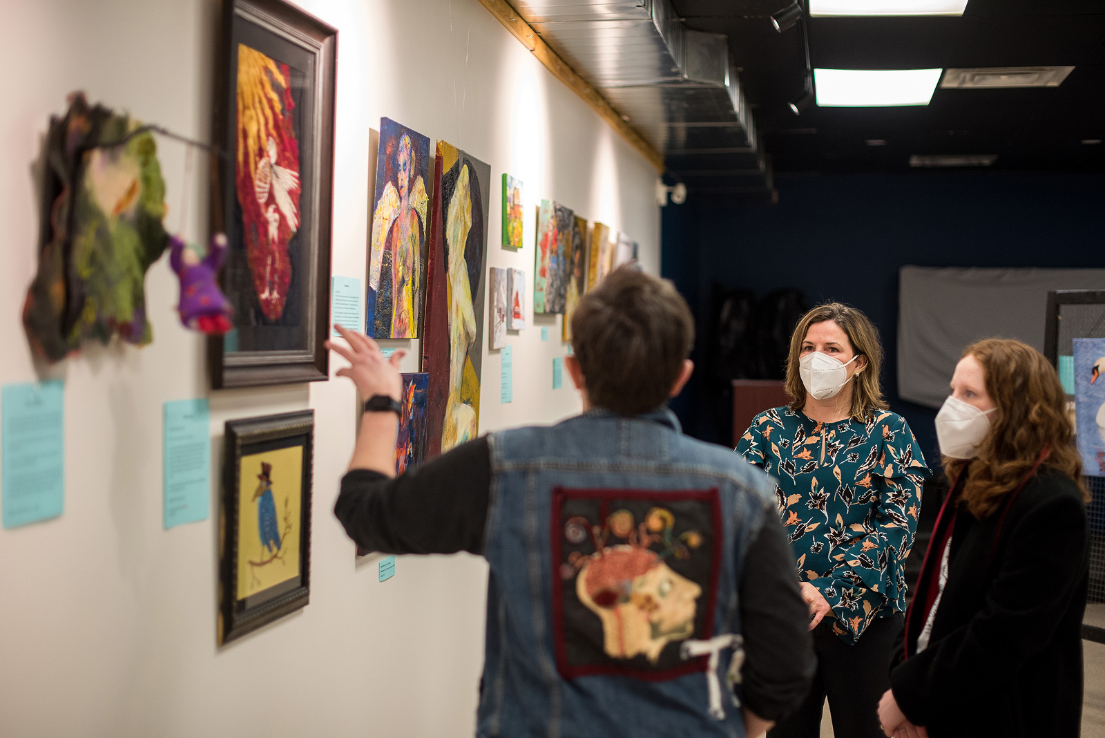Three people wearing masks are facing a wall of paintings at an art gallery.