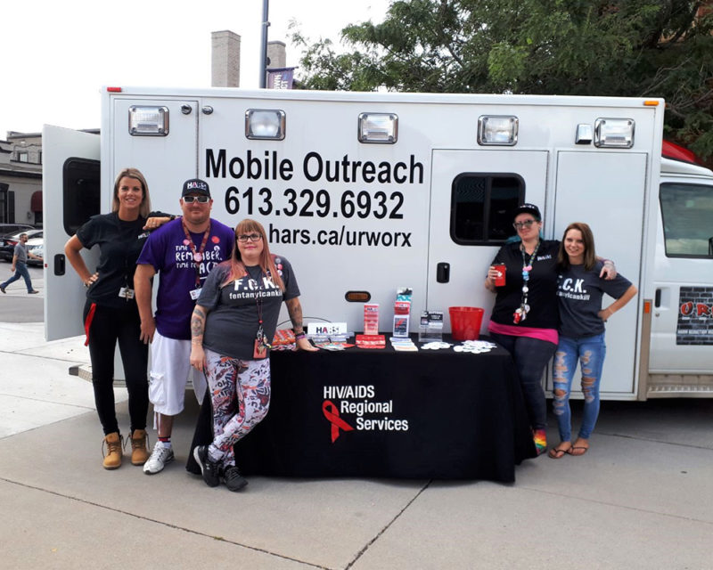 The Mobile Outreach truck. In front of the truck five team members are pictured alongside a table full of pamphlets.