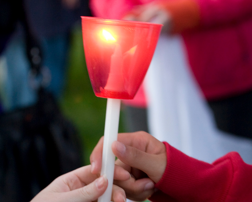 A red memorial candle is lit and being held by the hands of two people.