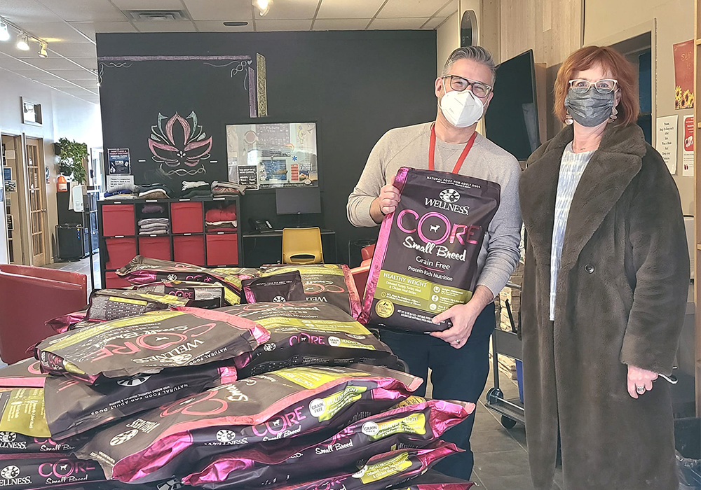 A man and woman standing indoors with masks standing beside a table covered in packages of dog food.