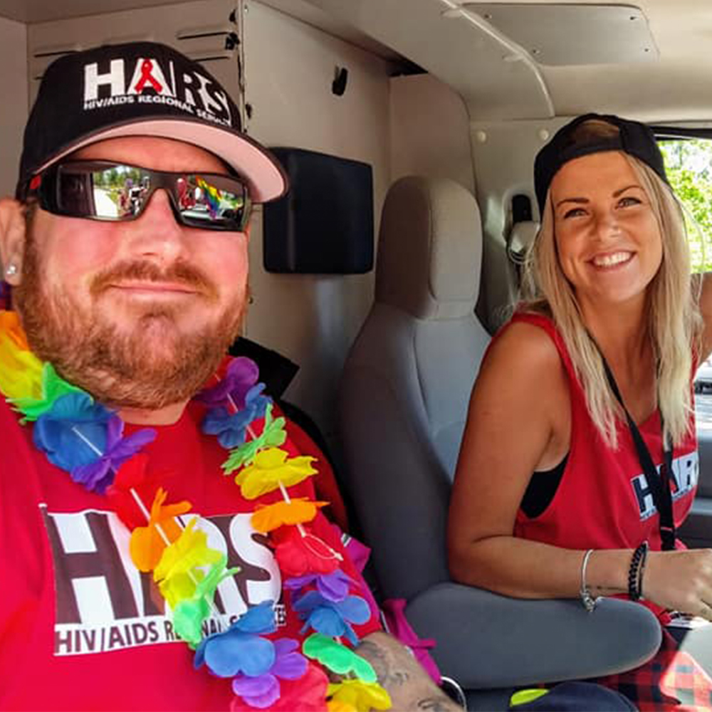 A man and a woman are facing the camera and smiling inside a car. They are both wearing red shirts and black hats with the HARS logo.