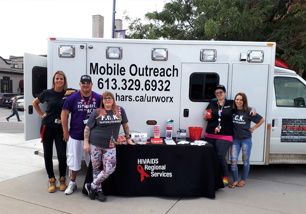 The Mobile Outreach truck. In front of the truck five team members are pictured alongside a table full of pamphlets.