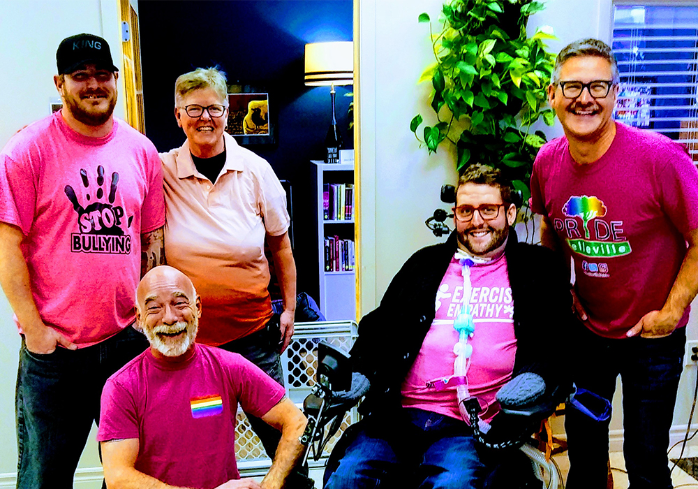 A group of 5 people, gathered posing for the camera. They are all wearing pink shirts and are smiling. One person is kneeling on the ground and one is a person who uses a wheelchair.