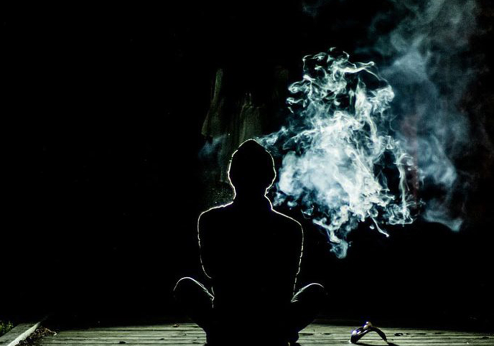 A person is sitting and facing away from the camera in a dark room. There is a cloud of white smoke emerging from the person.