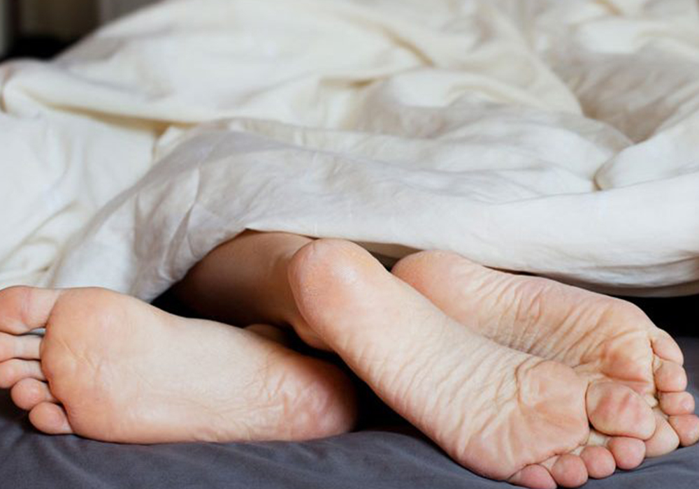 The feet of two people shown peaking out of a duvet cover in bed.